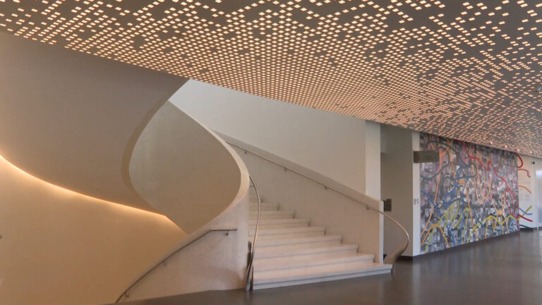 An elegant staircase inside a museum, highlighted by a patterned light ceiling and colorful abstract mural on the wall