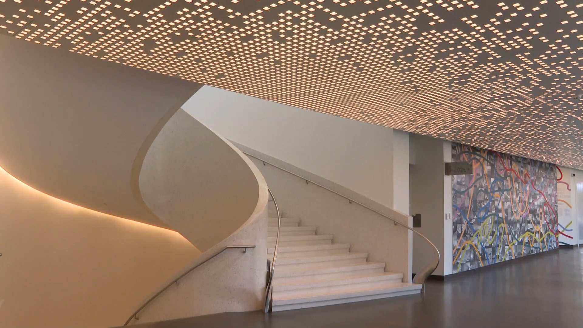 An elegant staircase inside a museum, highlighted by a patterned light ceiling and colorful abstract mural on the wall