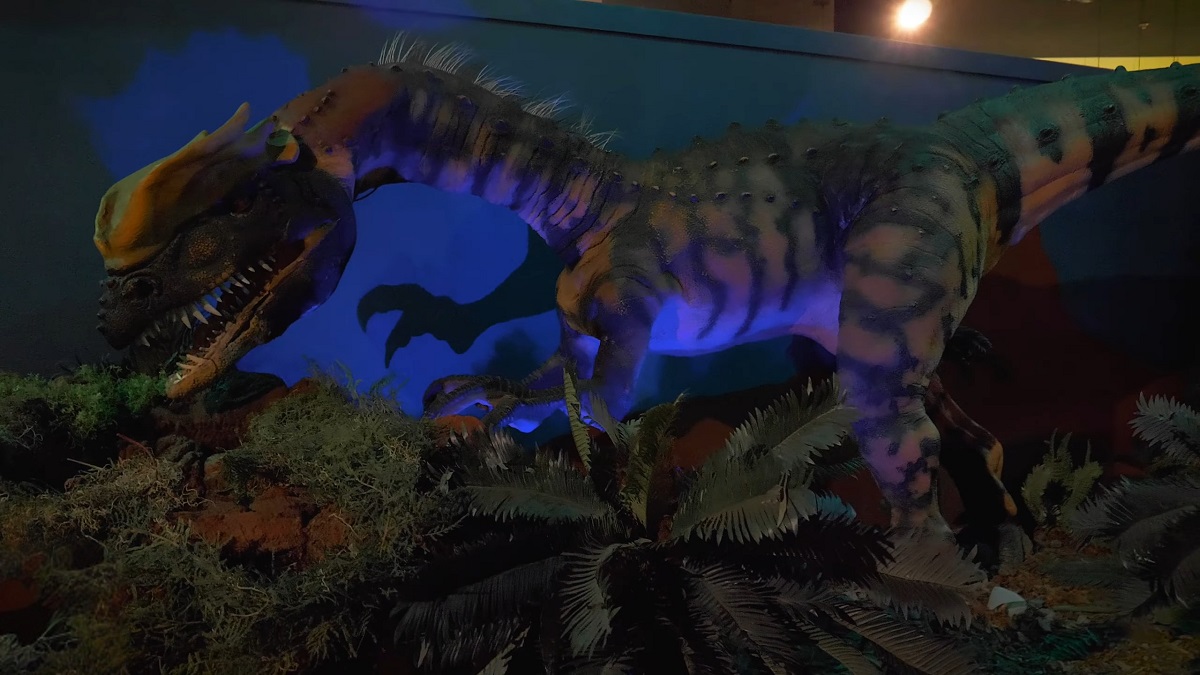 A vividly colored animatronic dinosaur with glowing eyes in a dark museum exhibit, surrounded by artificial plants and a blue-lit prehistoric setting